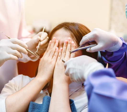 A patient is nervous at the dentist and hiding her face from the assistants, dentist, and dental tools.